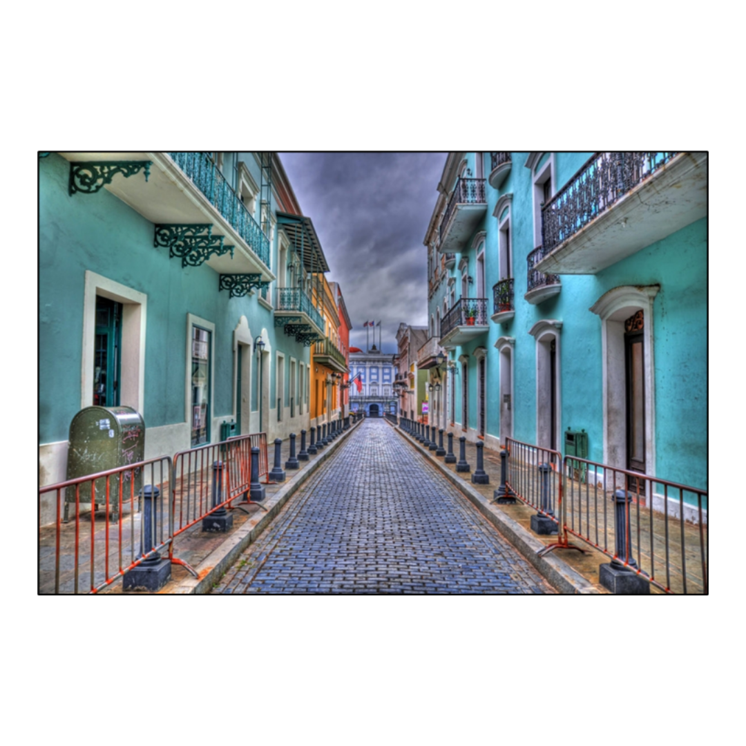 Brick street in San Juan lined on both sides with red barrier fences, curbed sidewalks and light blue buildings with ornate metal balconies.