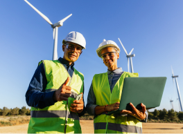 A clean energy worker mentoring a younger worker in a field of clean energy windmills