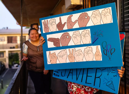 Hispanic people on a balcony hold up a sign showing hands forming sign language letters. 