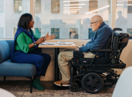 A woman interviews a man using a power wheelchair at a small table near a window.