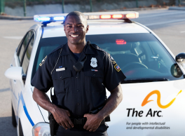 A police officer standing in front of his squad car. Logo: the Arc.