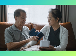 An elderly man sits next to his elderly wife on their couch and assists her in putting on a blood pressure cuff.