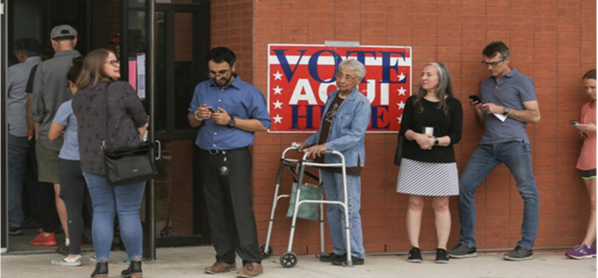 Una foto de varias personas esperando entrar a un edificio para votar. Hay un cartel grande con texto
	“Vote, Aquí, Here”, enfrente de el cartel una persona mayor utilizando una andadera.